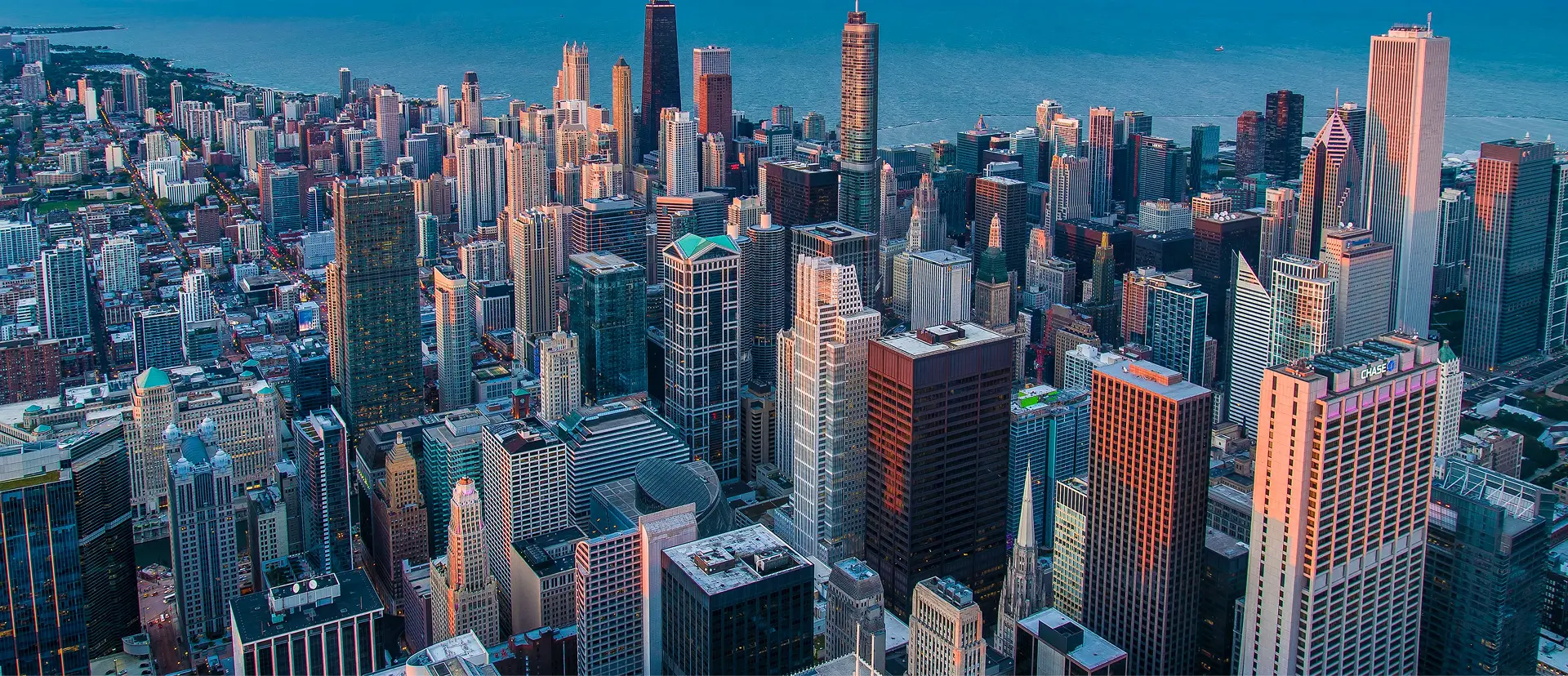 Aerial panoramic view of downtown Chicago skyline at dusk, showing Lake Michigan shoreline, high-rise buildings, the Chicago River area, and landmarks like the Tribune Tower and modern skyscrapers under a blue-purple sky.
